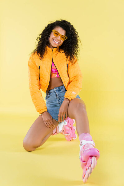 full length of smiling african american young woman in sunglasses and pink roller skates sitting on yellow