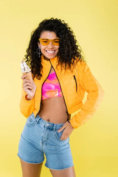 幸せなアフリカ系アメリカ人の若い女性ですサングラスをかけてアイスクリームコーンを黄色に隔離しました — ストック写真