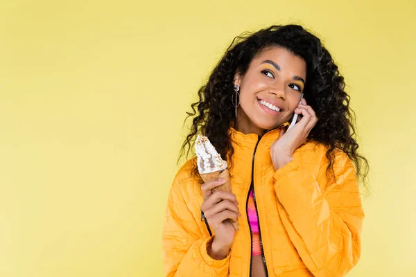 幸せなアフリカ系アメリカ人の若い女性がアイスクリームコーンを持っていて黄色で隔離された携帯電話で話しています — ストック写真