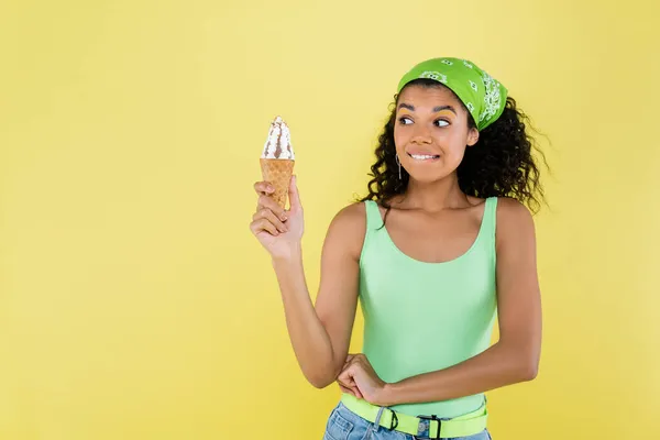 curly african american woman in green kerchief holding ice cream and biting lips isolated on yellow