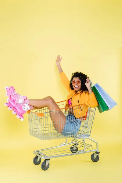 穿着粉色溜冰鞋 提着购物袋 坐在黄色购物车上的非洲裔美国年轻女子感到惊讶 — 图库照片