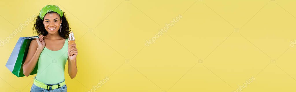 Happy african american woman in green kerchief holding ice cream cone and shopping bags isolated on yellow, banner