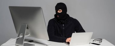 Bilgisayar yanında dizüstü bilgisayar ve akıllı telefon kullanan kar maskeli bilgisayar korsanı, gri pankartta izole edilmiş. 