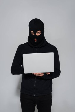 Siyah kar maskeli bir bilgisayar korsanı gri arka planda laptop kullanıyor. 