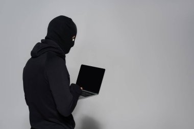 Kar maskeli bir bilgisayar korsanı gri arka planda boş ekranlı bir laptop kullanıyor. 