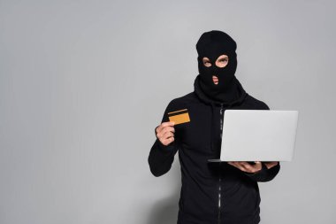 Kar maskeli bilgisayar korsanı gri arka planda kredi kartı ve laptop tutuyor. 