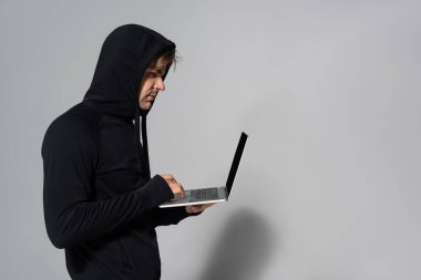 Gri arkaplanda dizüstü bilgisayar kullanan hacker 'ın yan görünümü 
