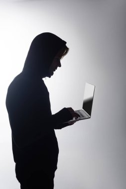 Gri ekranda izole edilmiş dizüstü bilgisayar kullanan başlıklı hacker yan görünümü 