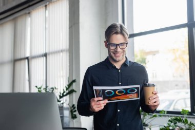 Ofisteki bilgisayarın yanına gitmek için elinde kağıt ve kahve tutan gülümseyen programcı. 