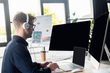 Masadaki kağıt bardağın yanında bilgisayar kullanan kulaklıklı programcının yan görüntüsü 