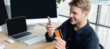Ofisteki bilgisayarların yanında akıllı telefon ve kredi kartı kullanan gülümseyen programcı, afiş 