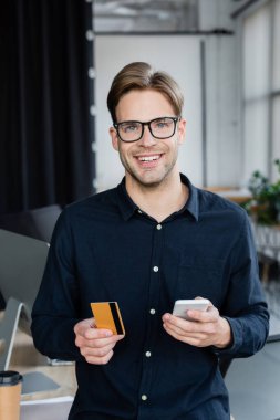 Ofisteki kameraya bakarken elinde kredi kartı ve cep telefonu tutan gülümseyen geliştirici. 