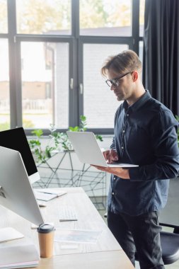 Gözlüklü genç programcı bilgisayarın yanında dizüstü bilgisayar ve ofiste kağıt bardak kullanıyor. 