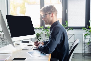 Ofiste boş ekranı ve akıllı telefonu olan bilgisayarların yanındaki deftere yazı yazan geliştiricinin yan görünümü 