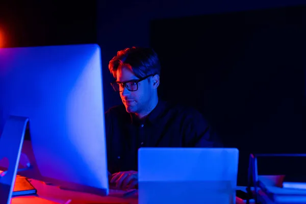 Young programmer in eyeglasses using computer near laptop on black background with lighting