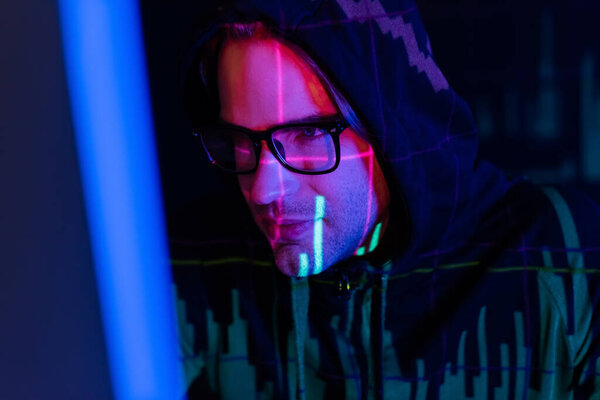 Young hacker in eyeglasses with charts reflection using blurred computer on black background 