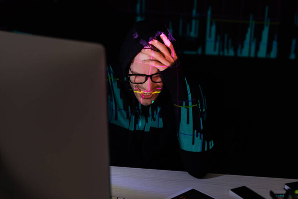 Sad hacker with reflection of charts sitting near devices on black background 