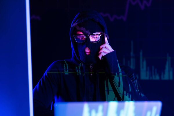 Pensive hacker in balaclava looking at laptop on black background with charts reflection 