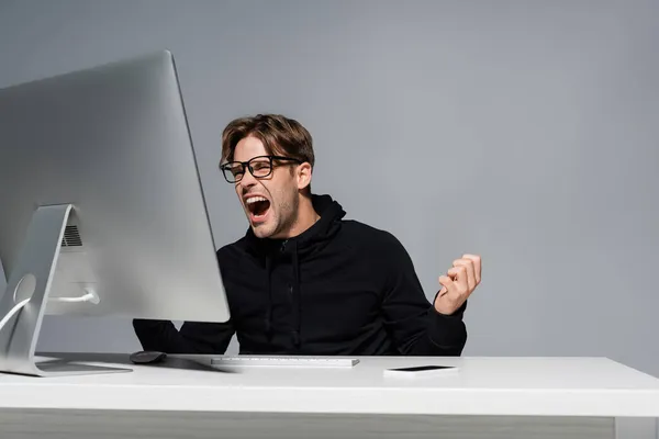 Angry programmer in eyeglasses screaming near computer and smartphone isolated on grey