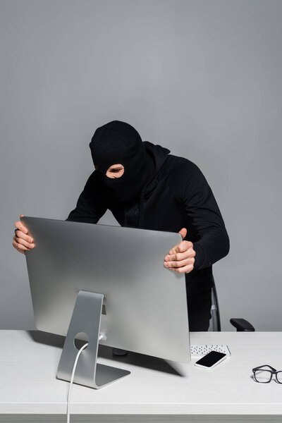Hacker in balaclava taking computer monitor near smartphone isolated on grey 