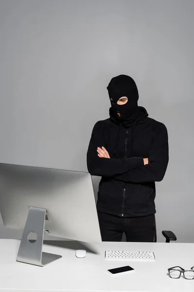 黑客身穿巴拉克拉瓦 双手交叉地站在电脑前 智能手机放在桌子上 与灰色隔离 — 图库照片