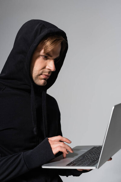 Young hacker using laptop while standing isolated on grey 