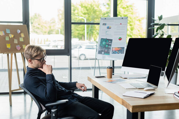 Pensive programmer looking at computers near smartphone and coffee on table 