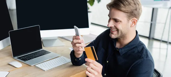 Uśmiechnięty Programista Pomocą Smartfona Karty Kredytowej Pobliżu Komputerów Biurze Baner — Zdjęcie stockowe