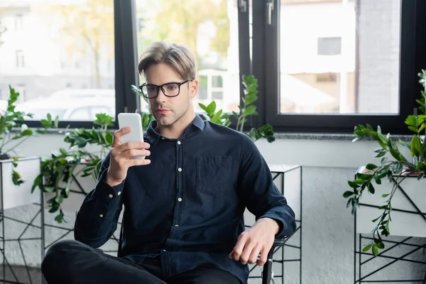 オフィスの椅子に座っている間 スマートフォンを使用して眼鏡の若い開発者 — ストック写真