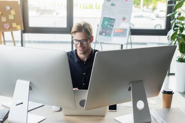 オフィスでノートパソコンやスマートフォンの近くのコンピュータを見てメガネの幸せな開発者 — ストック写真