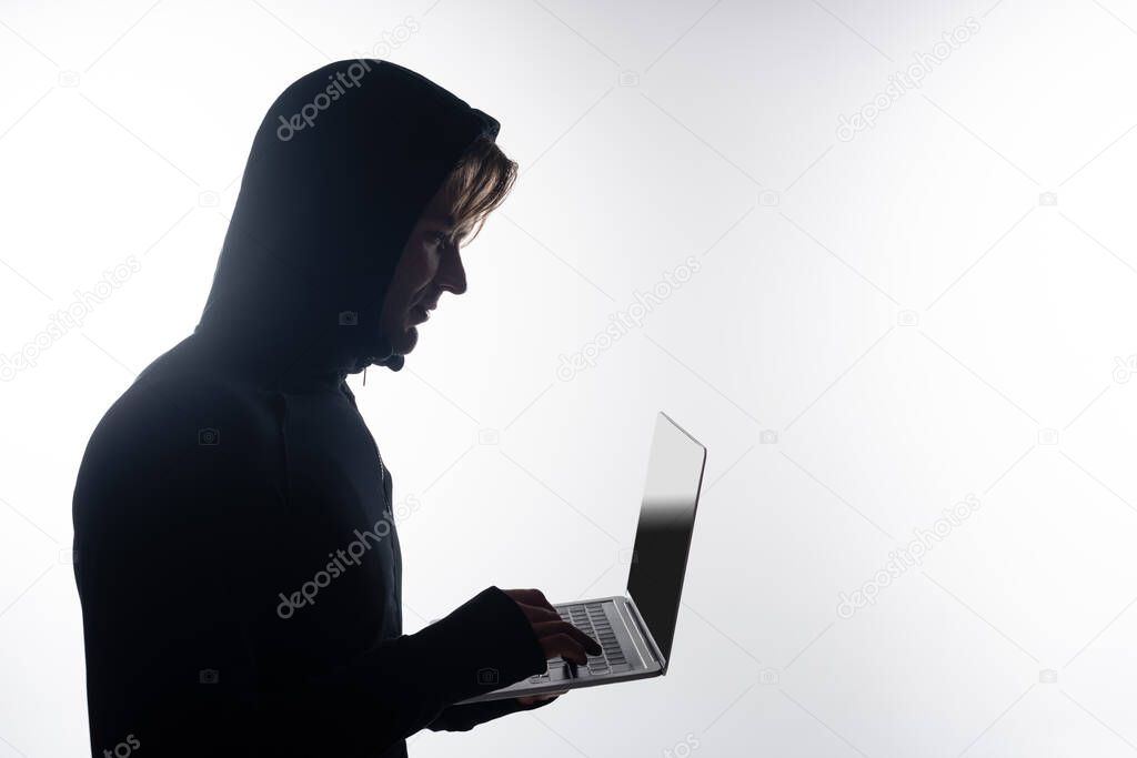 Side view of hacker in hood using laptop with blank screen isolated on grey 
