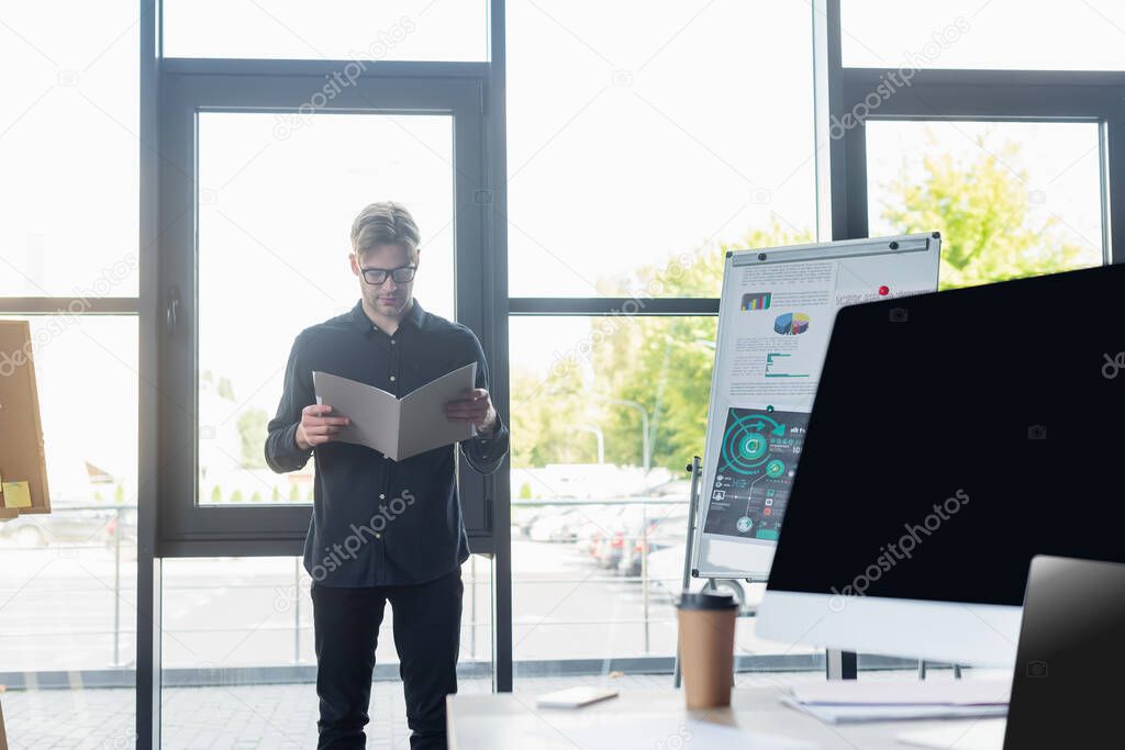 Programmer in eyeglasses holding paper folder near flip chart and devices in office 