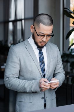 Resmi kıyafet ve gözlük giyen Asyalı işadamı ofisteki cep telefonundan mesaj yolluyor.