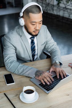 Kulaklıklı Asyalı işadamı laptopta kahve fincanı ve boş ekranlı akıllı telefonun yanında daktilo yazıyor.