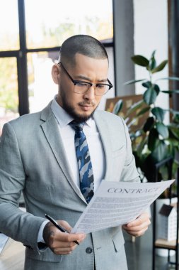 ciddi Asyalı iş adamı ofiste sözleşme okurken elinde kalem tutuyor.