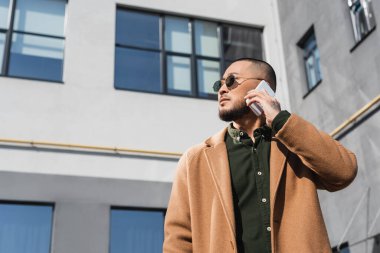 Asyalı güneş gözlüklü ve ceketli adamın düşük açılı görüntüsü şehirdeki akıllı telefondan arıyor.