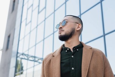 Güneş gözlüklü modaya uygun Asyalı adam cam cepheli bulanık binanın yakınına bakıyor.