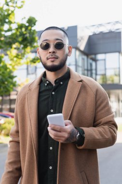 Havalı bir sonbahar kıyafeti ve güneş gözlüğü takan Asyalı bir adam dışarıda cep telefonu tutuyor.
