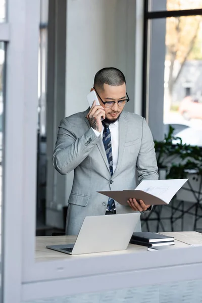 Asian Businessman Talking Mobile Phone While Standing Blueprint Laptop Desk — Stock Photo, Image
