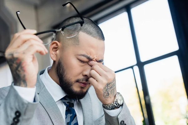 exhausted asian businessman rubbing eyes while holding blurred eyeglasses in office