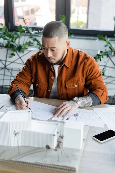 asian architect drawing blueprint while looking at blurred house models near smartphone with blank screen