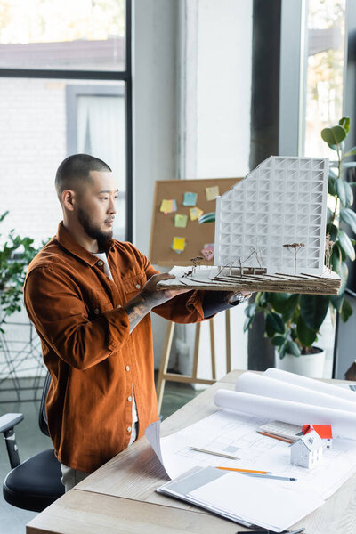 asian architect holding house model near blueprints on desk in office