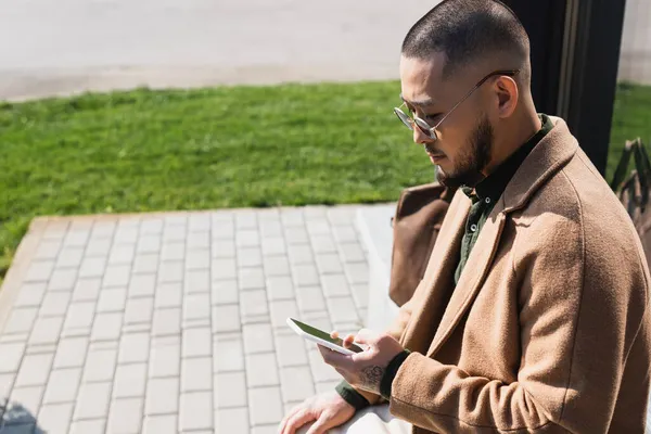 コートとサングラスを着たアジア系男性が屋外に座って携帯電話でメッセージを送り — ストック写真