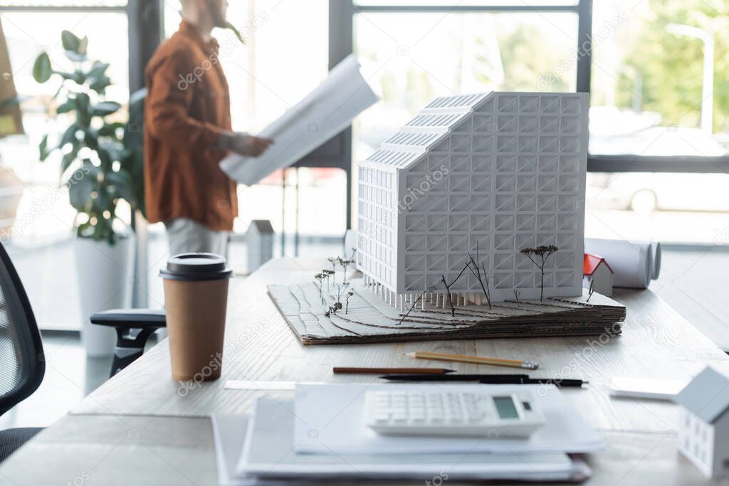 selective focus of house model and paper cup on desk near architect standing with paper on blurred background