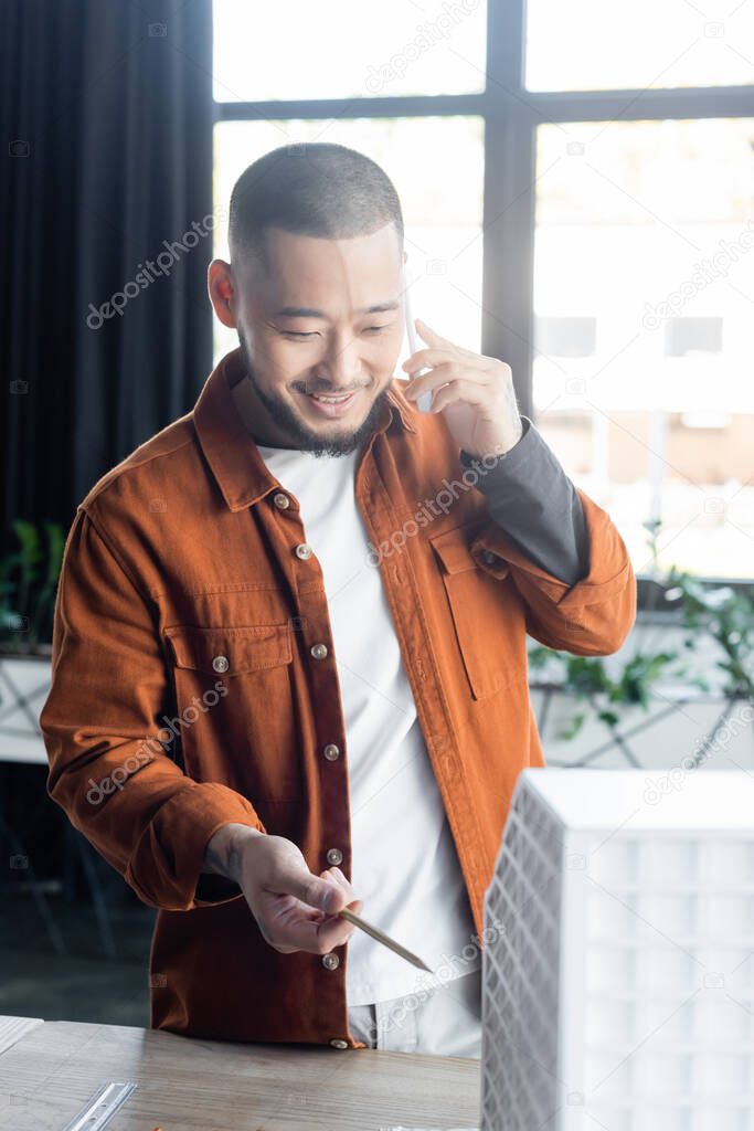 smiling asian architect talking on smartphone and pointing with pen at blurred house model 