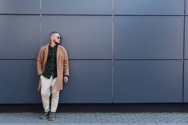 Gri duvarın yanında eli cebinde duran şık sonbahar kıyafetleri içindeki Asyalı adamın tam boy görüntüsü