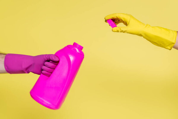 cropped view of people in rubber gloves holding pink bottle with detergent and bottle cap isolated on yellow 