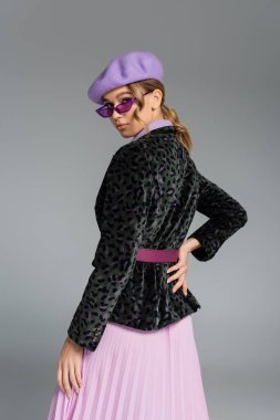 Bereli, spor ceketli, eli kalçasında poz veren gri elbiseli, modaya uygun genç bir kadın. 