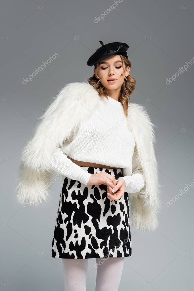 young woman in beret, white faux fur jacket and skirt with animal print posing isolated on grey