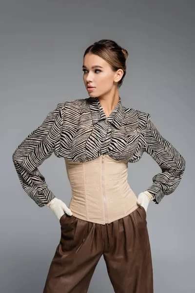 trendy woman in blouse with animal print, gloves and corset posing with hands in pockets isolated on grey